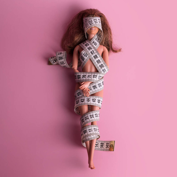 Doll wrapped in measuring tape. Tied up unrecognizable plastic doll, weight loss, fasting and slimming, diet, anorexia, overeating control, female fight for perfect fit body concept. Free copyspace - 写真・画像