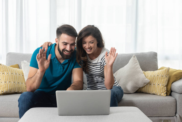Young couple sitting in their living room with laptop waving to their marital therapist after successful online therapy  and saved marriage   - 写真・画像