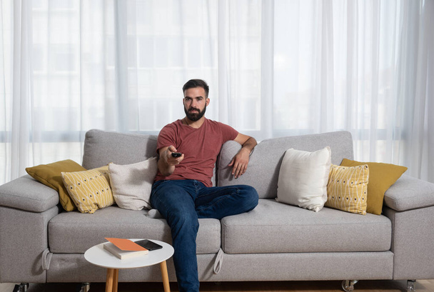 Young casual man sitting on the sofa at his home with remote control in his hand relaxing with television program - Foto, Bild