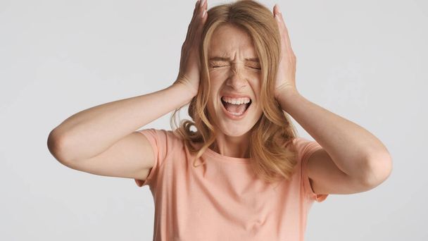Θλιβερό ξανθό κορίτσι που καλύπτει τα αυτιά συναισθηματικά ουρλιάζοντας στην κάμερα πάνω από λευκό φόντο - Φωτογραφία, εικόνα
