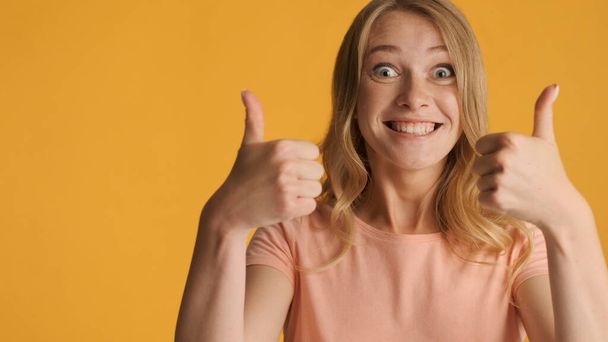 Excited smiling blond girl happily keeping thumbs up near with copy space for text or promotional content isolated on yellow background. Like expression - Photo, Image