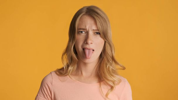 Fiatal, szomorú, szőke nő, aki színes háttérrel nyújtja ki a nyelvét a kamera előtt. Sértett kifejezés - Fotó, kép