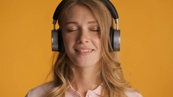 Beautiful dreamy blond girl listening music in headphones with eyes closed over yellow background - Photo, Image