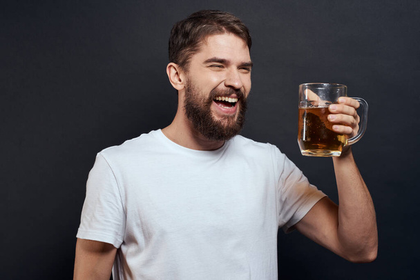 Mężczyzna z kubkiem piwa w rękach emocje zabawa styl życia biały t-shirt ciemne odizolowane tło - Zdjęcie, obraz