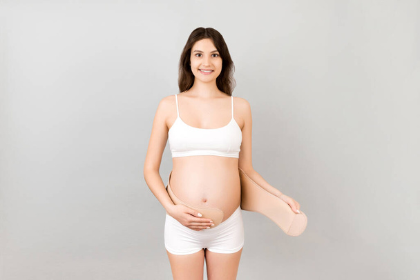 Portrait of pregnant woman in white underwear putting on a bandage at gray background with copy space. Orthopedic abdominal support belt concept. - Photo, Image