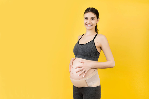 Ritratto di donna incinta che indossa un corsetto di gravidanza contro il mal di schiena su sfondo giallo con spazio per la copia. Cintura di supporto addominale ortopedico concetto. - Foto, immagini