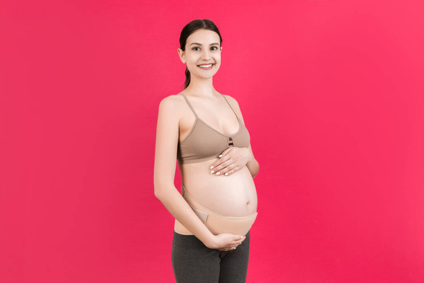 Portrait of pregnant woman wearing pregnancy corset against backpain at pink background with copy space. Orthopedic abdominal support belt concept. - 写真・画像