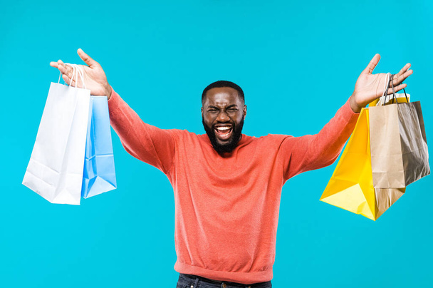 Gros achats. Heureux homme afro-américain debout sur fond bleu tenant des sacs à provisions en papier souriant et regardant la caméra - Photo, image