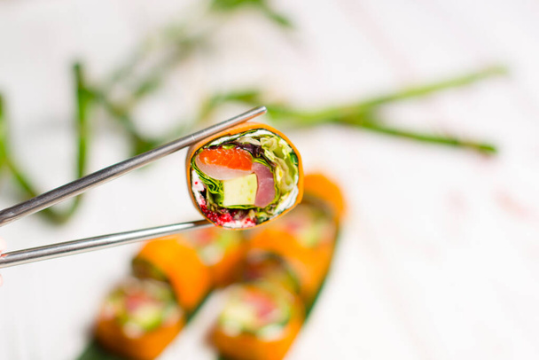 Close up de Sushi Roll pedaço sem arroz com abacate, salmão, atum, filé de rockfish envolto em daikon cenoura em pauzinhos de metal com fundo branco desfocado de folhas de bambu e planta. Sem arroz - Foto, Imagem