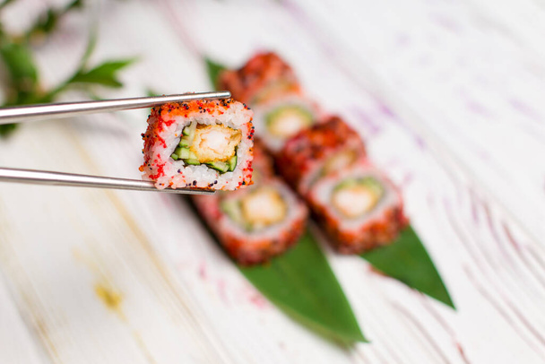 Detailní záběr Sushi Roll kus zabalený v rýži a létající ryby jikry v kovových hůlkách s rozmazaným pozadím. Sushi Roll s tempura krevety uvnitř na bambusových listech a rostliny na pozadí. Kopírovat prostor - Fotografie, Obrázek