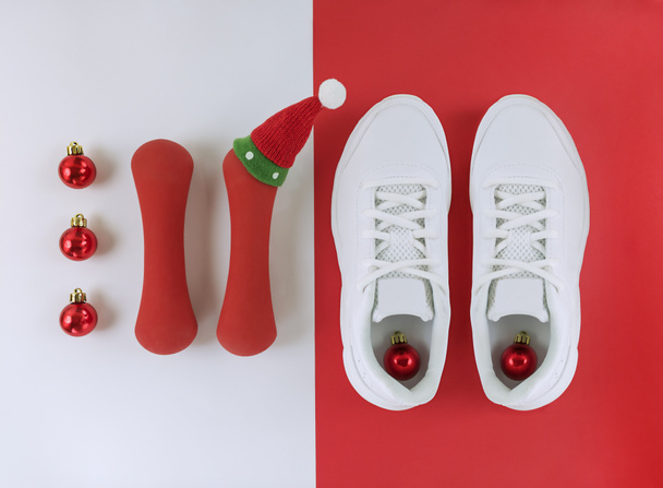 Sport holiday flat lay. Christmas balls, trainers, and dumbbells with cute hat on white and red background. - Photo, Image
