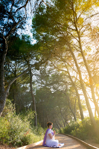 Mulher meditando sentada no lótus posar com as mãos na frente de seu peito, em anjali mudra, em um caminho florestal ao pôr do sol - Foto, Imagem