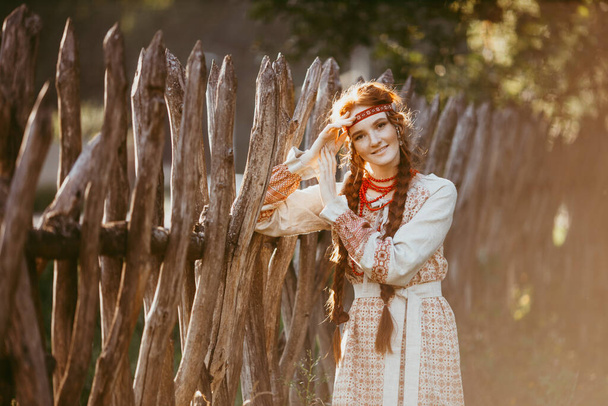 A beautiful Slavic girl with long blonde hair and brown eyes in a white and red embroidered suit stands by a wooden fence.Traditional clothing of the Ukrainian region.summer day - Φωτογραφία, εικόνα