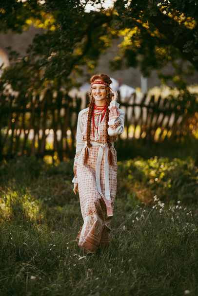 A beautiful Slavic girl with long blonde hair and brown eyes in a white and red embroidered suit stands by a wooden fence.Traditional clothing of the Ukrainian region.summer day - Photo, image