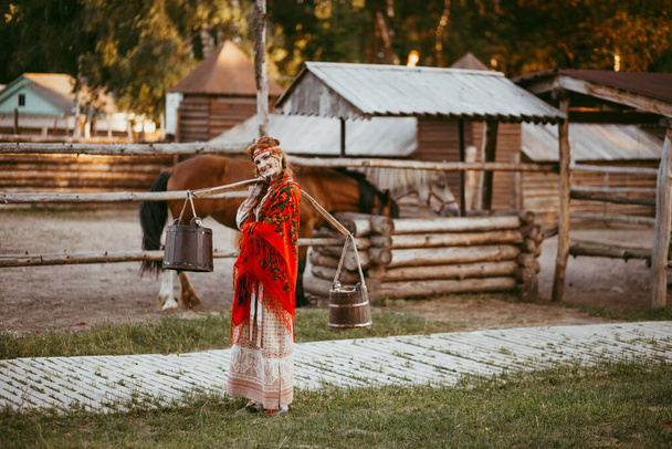 A beautiful Slavic girl with long blonde hair and brown eyes in a white and red embroidered suit is sitting next to a wooden house.Traditional clothing of the Ukrainian region.summer day - Φωτογραφία, εικόνα