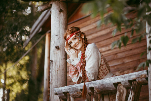 A beautiful Slavic girl with long blonde hair and brown eyes in a white and red embroidered suit stands on the porch of a house.Traditional clothing of the Ukrainian region.summer day - Photo, image