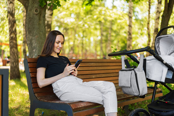 молодая мама, женщина со смартфоном в руках сидит на скамейке рядом с коляской с ребенком, счастливая долгожданная концепция материнства - Фото, изображение