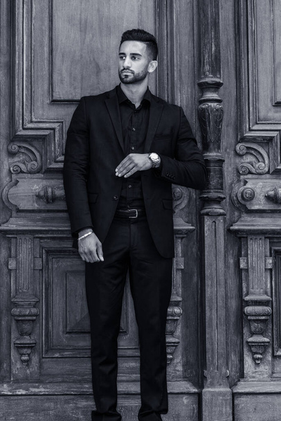 Young East Indian American Businessman with beard, wearing black suit, wristwatch, standing by old style office doorway in New York City, hand in front touching clothes, looking away. Black and White - Photo, Image
