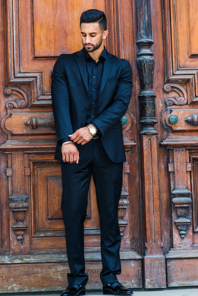 若い東インド系アメリカ人のビジネスマン、ひげ、黒のスーツを着て、革の靴、腕時計、ニューヨークの茶色の古いスタイルのオフィスの入り口で立って、手をまっすぐに袖 - 写真・画像