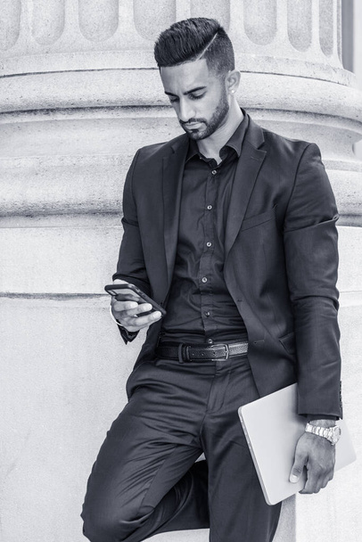 A fiatal kelet-indiai amerikai üzletember szakállal New Yorkban dolgozik, fekete öltönyt visel, laptopot cipel, irodaépület előtt áll, mobilon SMS-ezik. Fekete-fehér - Fotó, kép