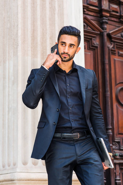 Young East Indian American Businessman traveling, working in New York City, with beard, wearing black suit, carrying laptop computer, walking from old style office doorway, talking on cell phone - Fotó, kép