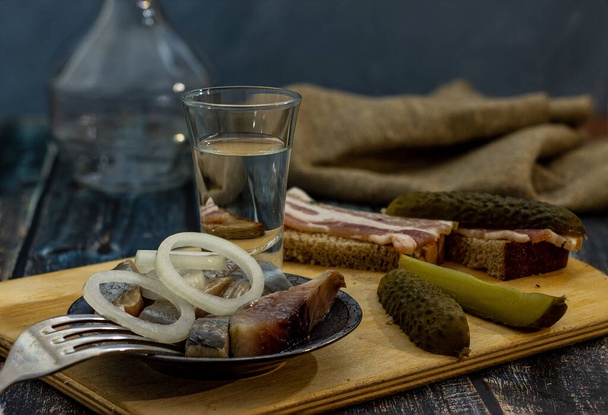 Стакан алкоголю та закуски: оселедце з цибулею на тарілці, огірок, бекон та хліб
. - Фото, зображення