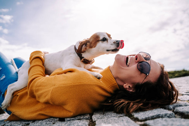 A beautiful woman laughing while her pet is licking her face in a sunny day in the park in Madrid. The dog is on its owner between her hands. Family dog outdoor lifestyle - Photo, Image