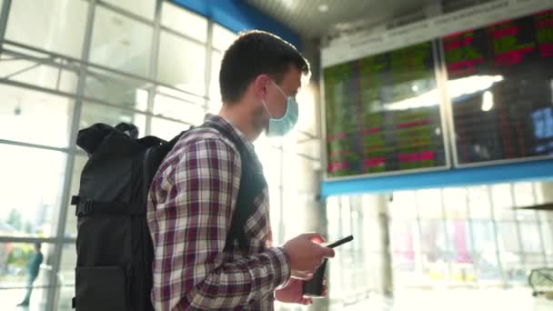 Hombre con máscara protectora y taza de café usar el teléfono inteligente para el check-in en línea de pie en la terminal del aeropuerto cerca de la salida horario de llegada pantalla durante la cuarentena covid 19, virus corona - Metraje, vídeo