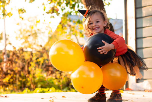 Una linda niña 2-3 en un traje de bruja de Halloween naranja y negro con globos, de pie en la terraza de una casa gris de madera - Foto, imagen