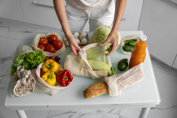 Woman came back from a market and unpacks a reusable grocery bag full of vegetables on a kitchen at home. Zero waste and plastic free concept. Girl is holding mesh cotton shopper with vegetables. - Foto, Imagen