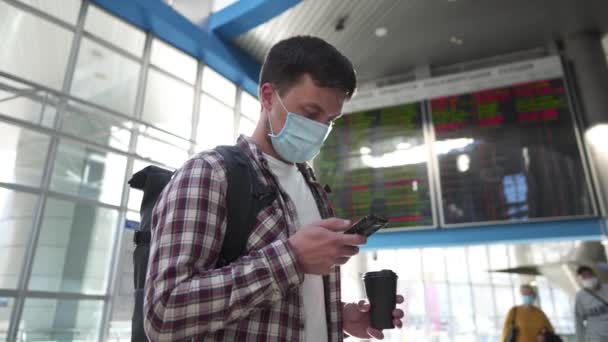 Man in protective mask and cup coffee use smart phone for online check-in standing in airport terminal near departure arrival timetable screen display during quarantine covid 19, corona virus - Footage, Video