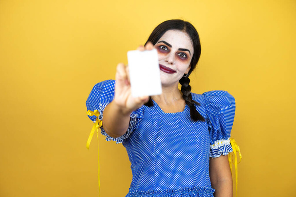 mulher vestindo uma fantasia de Halloween boneca assustadora sobre fundo amarelo sorrindo e segurando cartão branco - Foto, Imagem