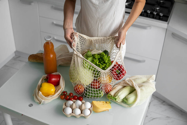 Woman came back from a market and unpacks a reusable grocery bag full of vegetables on a kitchen at home. Zero waste and plastic free concept. Girl is holding mesh cotton shopper with vegetables. - Fotografie, Obrázek