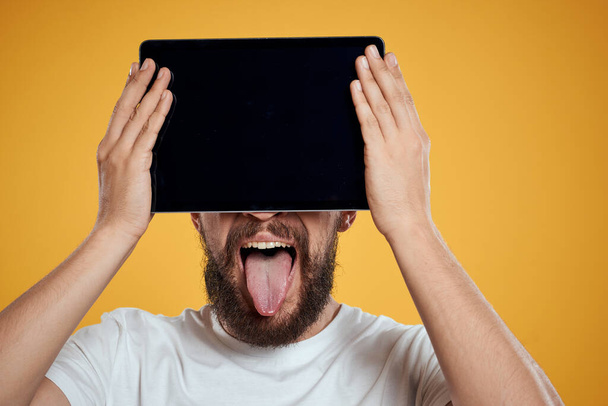 Portret van een man met een tablet voor zijn gezicht op een gele achtergrond bijgesneden weergave van een wit T-shirt Copy Space - Foto, afbeelding