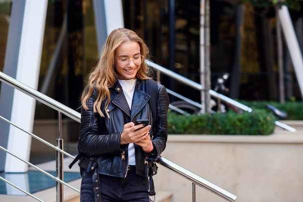 Cheerful beautiful student girl uses smart phone at stairs on the street, enjoying walk outdoors. - Zdjęcie, obraz