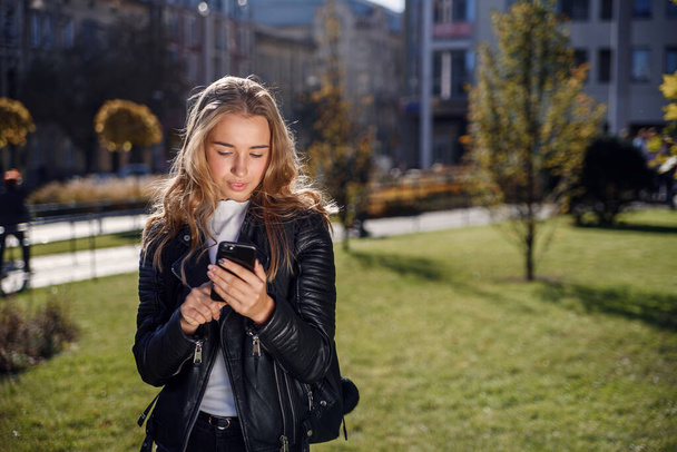 Beautiful girl writing or reading messages online on a smart phone while walking on the street at sunny day. - Foto, imagen