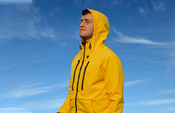 ελκυστικός μεσήλικας άνδρας σε ζωηρό κίτρινο αδιάβροχο αναζητούν τρόπους πλευρά με ελαφρώς χαμόγελο σε μπλε ουρανό άσπρα σύννεφα φόντο εξωτερική γραφική θέα και κενό χώρο αντίγραφο για το κείμενό σας εδώ - Φωτογραφία, εικόνα