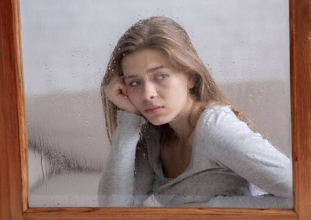 Smutna młoda kobieta siedząca w domu i wyglądająca przez okno na padający deszcz, cierpiąca na jesienną depresję - Zdjęcie, obraz
