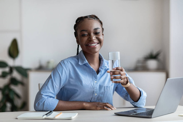Hidratación en el trabajo. Feliz negro mujer oficina trabajador sosteniendo vaso de agua - Foto, imagen