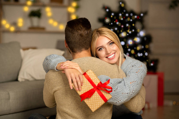 Задоволена дружина тримає різдвяний подарунок і обіймає чоловіка
 - Фото, зображення