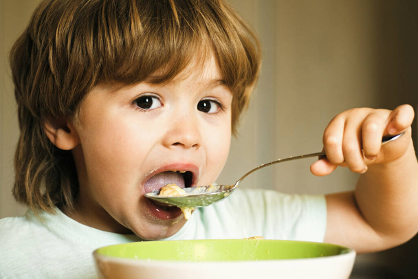 Hungry little boy eating. Cheerful baby child eats food itself with spoon. Tasty kids breakfast. Baby eating food on kitchen. Happy baby boy eats healthy food spoon itself. - Photo, image