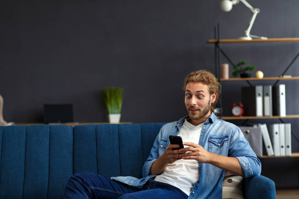 Curly-haired guy in casual style, watching great news on his mobile phone. Student receiving sms message reading good news. Surprised Man celebrating Victory on phone in Apartment - Photo, Image