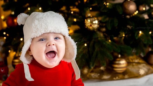 Jouluvauva hymyilee. Söpö pieni tyttö punaisessa mekossa ja valkoisessa hatussa ilmaisee tunteita. Joulukonsepti pieni lapsi, puu ja seppele taustalla hämärä - Valokuva, kuva