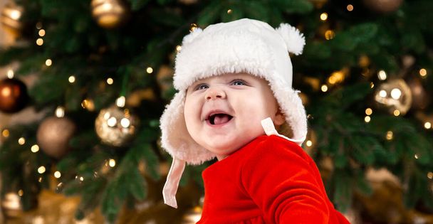 Vánoční dítě se usmívá. Roztomilá holčička v červených šatech a bílém klobouku vyjadřuje emoce. Vánoční koncept s malým dítětem, strom a girland na pozadí v rozmazání - Fotografie, Obrázek
