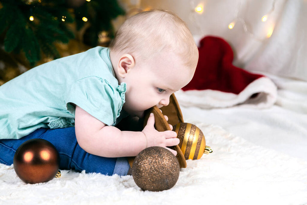 il bimbo in jeans e scarpe si siede vicino a un albero di Natale, le palle di giocattoli si trovano intorno. Bambino gioca con la gamba. - Foto, immagini