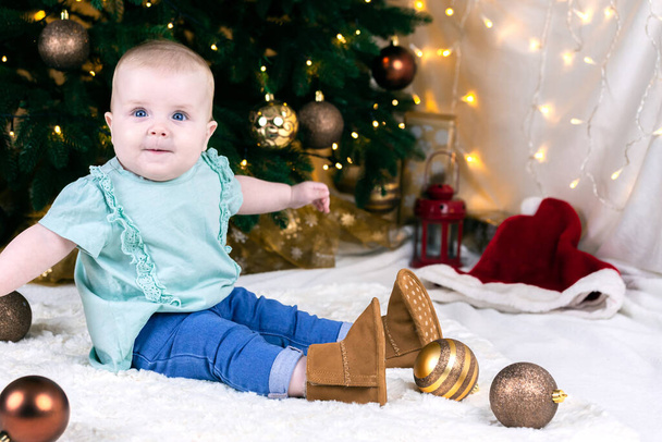 Kotlu ve ayakkabılı bebek Noel ağacının yanında oturuyor, oyuncak toplar etrafta yatıyor..  - Fotoğraf, Görsel