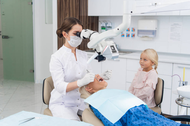 Bir dişçi, özel bir tıbbi aletin yardımıyla dişçi koltuğunda hastasının ağız boşluğunu inceler. - Fotoğraf, Görsel