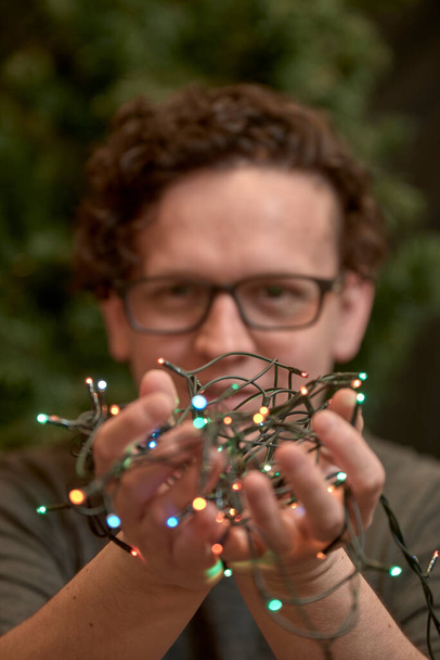 Young caucasian man with glasses holding a string of colored Christmas lights in both hands. Selective focus. - Zdjęcie, obraz