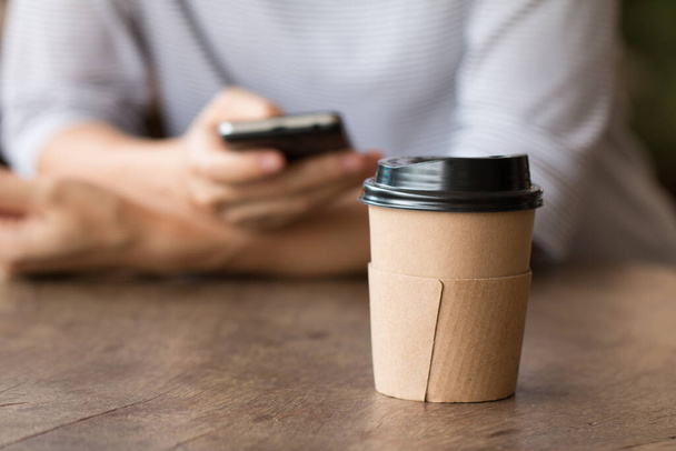 Taza de café de papel bebida caliente en el escritorio de madera con la mujer que trabaja en el teléfono inteligente borrosa en el fondo. - Foto, imagen