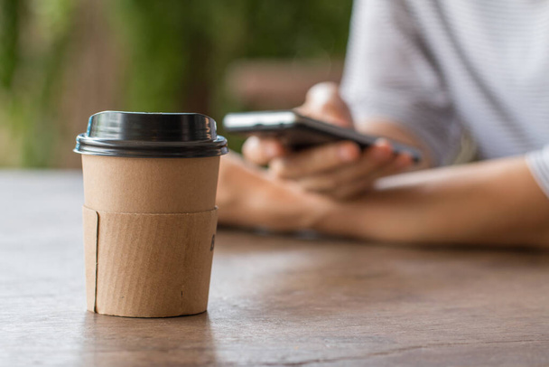 Papieren koffiekopje warme drank op houten bureau met vrouw die werkt op smart-phone wazig op de achtergrond. - Foto, afbeelding